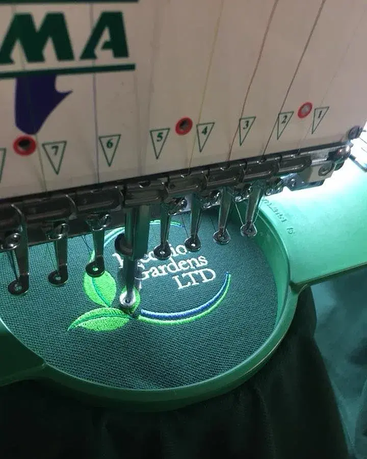 Nadelleiste der Stickmaschine über einem Stickrahmen mit grüner Logostickerei